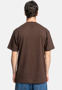 Camiseta de algodón marrón con un corte relajado, mangas cortas y una parte posterior lisa. Presenta un sutil logo cerca del escote.