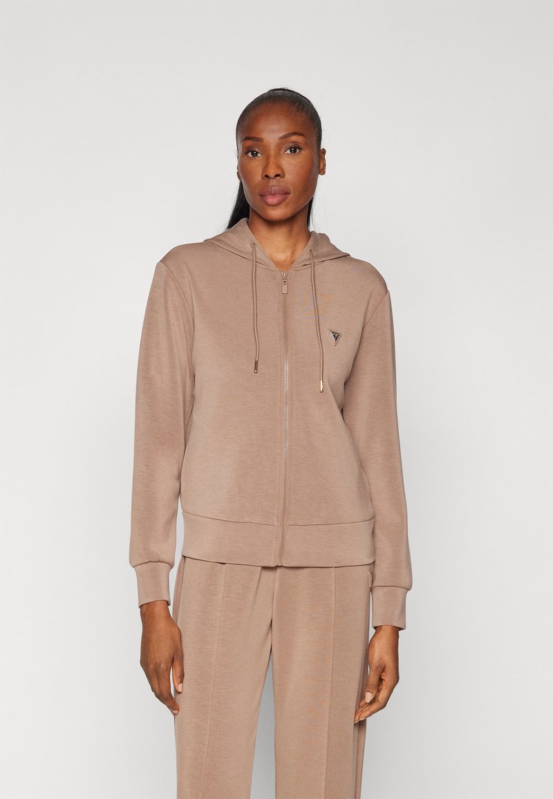 Felpa con zip beige realizzata in tessuto morbido, con cappuccio, polsini a costine e un piccolo logo sul petto. Abbinata a pantaloni coordinati.