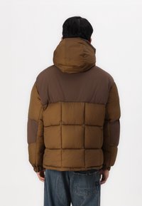 Braune Pufferjacke mit Kapuze, ausgestattet mit einem gesteppten Design, kontrastierendem braunem Oberteil und elastischen Bündchen. Trägt dazu weite blaue Jeans.