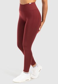 Kvinna klädd i högmidjade burgunderfärgade leggings och vita sneakers med märkesstrumpor, stående mot en slät ljus bakgrund.