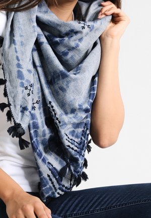 Femme portant une grande écharpe bleue tie-dye avec des pompons noirs et une broderie florale par-dessus une chemise blanche et un jean foncé.