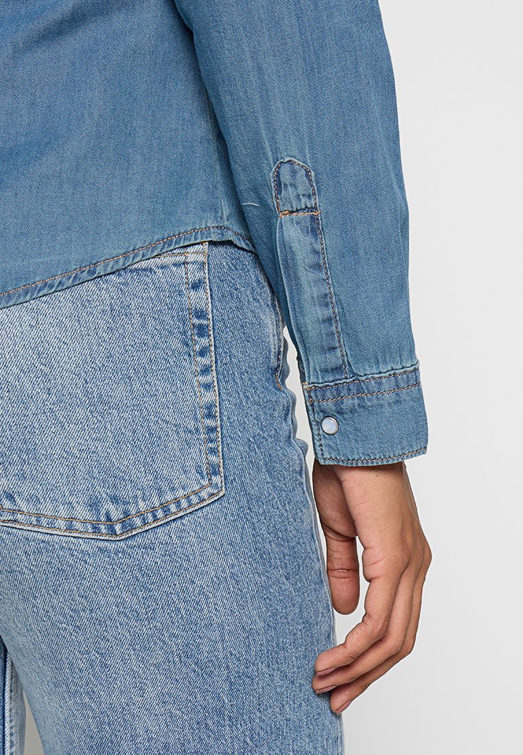 Veste en denim bleu clair avec des coutures contrastantes, détail des boutons aux poignets et une coupe décontractée. Associée à un jean bleu clair avec une poche arrière.
