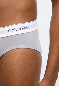 Sivé bavlnené nohavičky s bielym elastickým pásom s logom "Calvin Klein" v čiernej farbe. Hladká textúra, priliehavý strih a minimalistický dizajn.