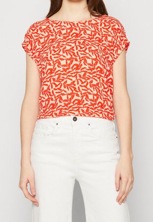 Kobieta ubrana w krótki czerwono-biały top w liściasty wzór, zestawiony z białymi, wysokim stanem jeansami, na jednolitym tle.