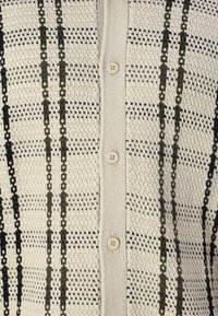 Strikket cardigan med en kremfarget bakgrunn og svart og olivengrønn rutemønster. Fem knapper foran, teksturert stoff.