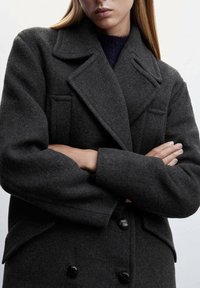 Donkergrijze wollen jas met een dubbele knoopsluiting, ingezette klappen en knoopaccenten. De mouwmanchetten hebben een gevouwen textuur.