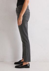 Graue Denim-Jeans mit geradem Schnitt, ausgestattet mit Gesäßtaschen und einem Reißverschluss. Kombiniert mit glänzenden schwarzen Loafers.