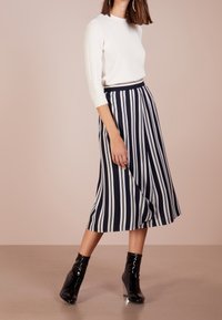 Donna che indossa una maglietta bianca a maniche lunghe, una gonna midi a strisce verticali blu scuro e bianche, e stivaletti neri lucidi, in piedi su un pavimento beige.