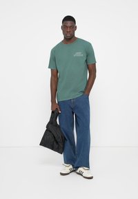 Grøn kortærmet t-shirt med et logo, parret med blå vide jeans og hvide sneakers med sorte striber. Holder en sort jakke.