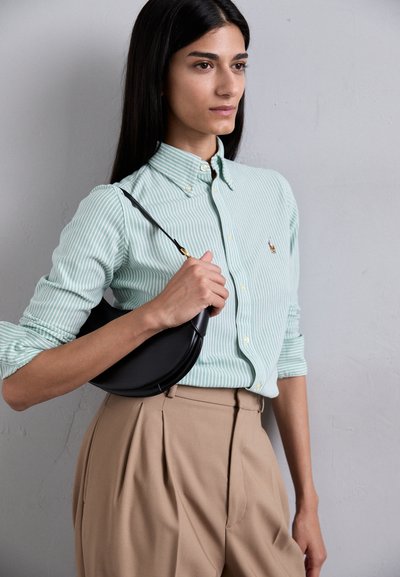 Camicia a righe verde chiaro con bottoni e pantaloni beige a vita alta. Borsa a forma di mezzaluna nera con tracolla, tenuta in una mano.