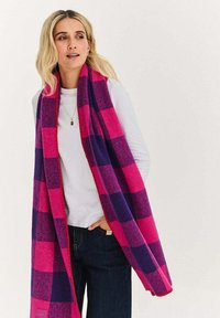Wełniany szal w mieszance włókien, ozdobiony wyrazistym wzorem kratki w fioletowo-różowych odcieniach z fakturowanym wykończeniem. Krawędzie obszyte kontrastującym czerwonym nicią.
