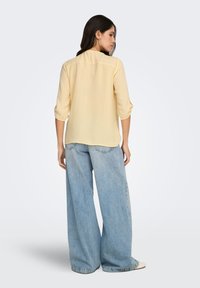 Lichtgeel overhemd met lange mouwen en omgeslagen manchetten; losse pasvorm. Gecombineerd met lichtblauwe, wijde jeans met een ontspannen silhouet.