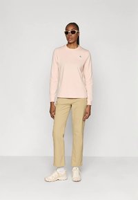 Mulher vestindo uma camisa de mangas longas rosa clara, calças bege, tênis creme e óculos de sol com armação branca, de pé contra um fundo claro e simples.