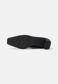 Zapato plano negro con suela de goma texturizada, tacón rectangular y patrón de huella geométrica. Material superior liso sin logotipos visibles.