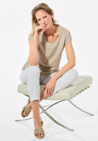 Beige Kurzarmoberteil mit Rundhalsausschnitt, kombiniert mit weißen Slim-Fit-Hosen und gewebten beigen Sandalen, sitzt auf einer hellen Bank.