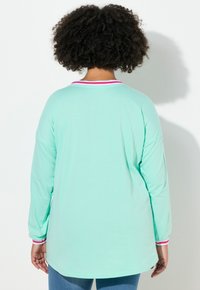 Persona con capelli ricci che indossa una camicia a maniche lunghe blu chiaro con colletto e polsini a righe rosa e bianche, di spalle rispetto alla fotocamera.