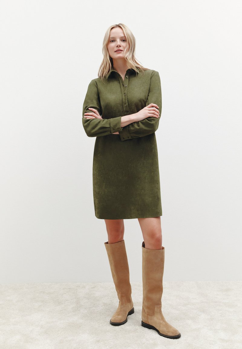Olivengrünes Hemdkleid mit einem geknöpften Kragen, langen Ärmeln und einer weichen Textur. Kombiniert mit hohen beigen Wildlederstiefeln und einem glatten Teppichhintergrund.