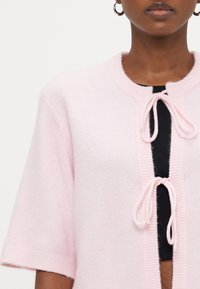 Pink knitted cardigan featuring short sleeves, a round neckline, and a front tie closure. Soft texture with ribbed accents along the edges.