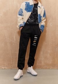 Persona che indossa una giacca di pile bianca e blu a motivi, una maglietta nera con grafica, pantaloni tuta neri Nike con più loghi e scarpe bianche.