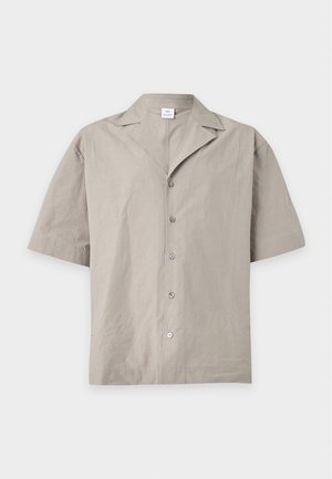 Joven de pie con las manos en los bolsillos, vistiendo una camisa beige de manga corta, vaqueros de pierna ancha azul claro y zapatos negros sobre fondo blanco.