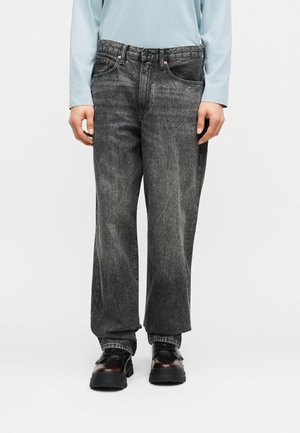 Isik, kes kannab lahtisi tuhmunud musta värvi teksapükse, mustade massiivsete jalanõudega ja helesinist pika varrukaga pluusi, seisab valge tausta ees.