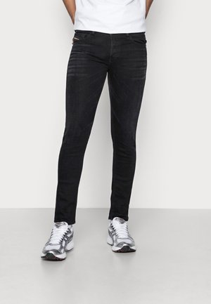 Homme portant un jean noir slim et des chaussures de course blanches et grises, debout sur un sol uni gris clair avec les mains dans les poches.