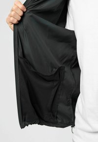 Käsi hoiab lahtist musta jopet, näidates sisemist võrgust taskut ja siledat voodrit valge särgi taustal.