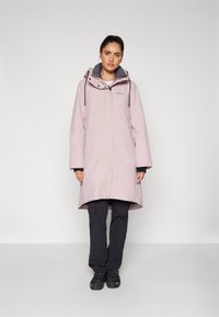 Cappotto impermeabile rosa chiaro con cappuccio, zip anteriore e foderato in grigio. Abbinato a pantaloni scuri e stivali neri, esposto su uno sfondo semplice.