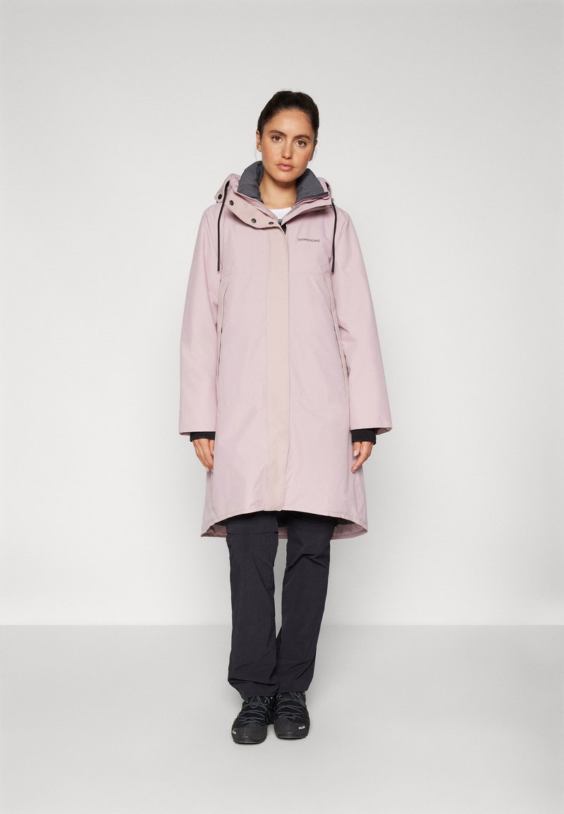 Cappotto impermeabile rosa chiaro con cappuccio, zip anteriore e foderato in grigio. Abbinato a pantaloni scuri e stivali neri, esposto su uno sfondo semplice.