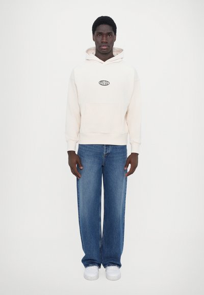 Krämfärgad luvtröja med framficka och logotyp, kombinerad med lösa blå denimjeans och vita sneakers, på en neutral bakgrund.
