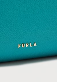 Τσάντα από δέρμα σε τιρκουάζ χρώμα με υφή, που διαθέτει χαραγμένο χρυσό λογότυπο "FURLA" σε εμφανές σημείο. Συμπαγής ορθογώνιο σχήμα.