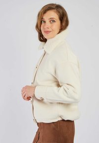 Kremowa wełniana kurtka z miękkim kołnierzem wyściełanym polarem, zapinana na guziki, z ściągaczami na rękawach, połączona z brązową zamszową spódnicą.