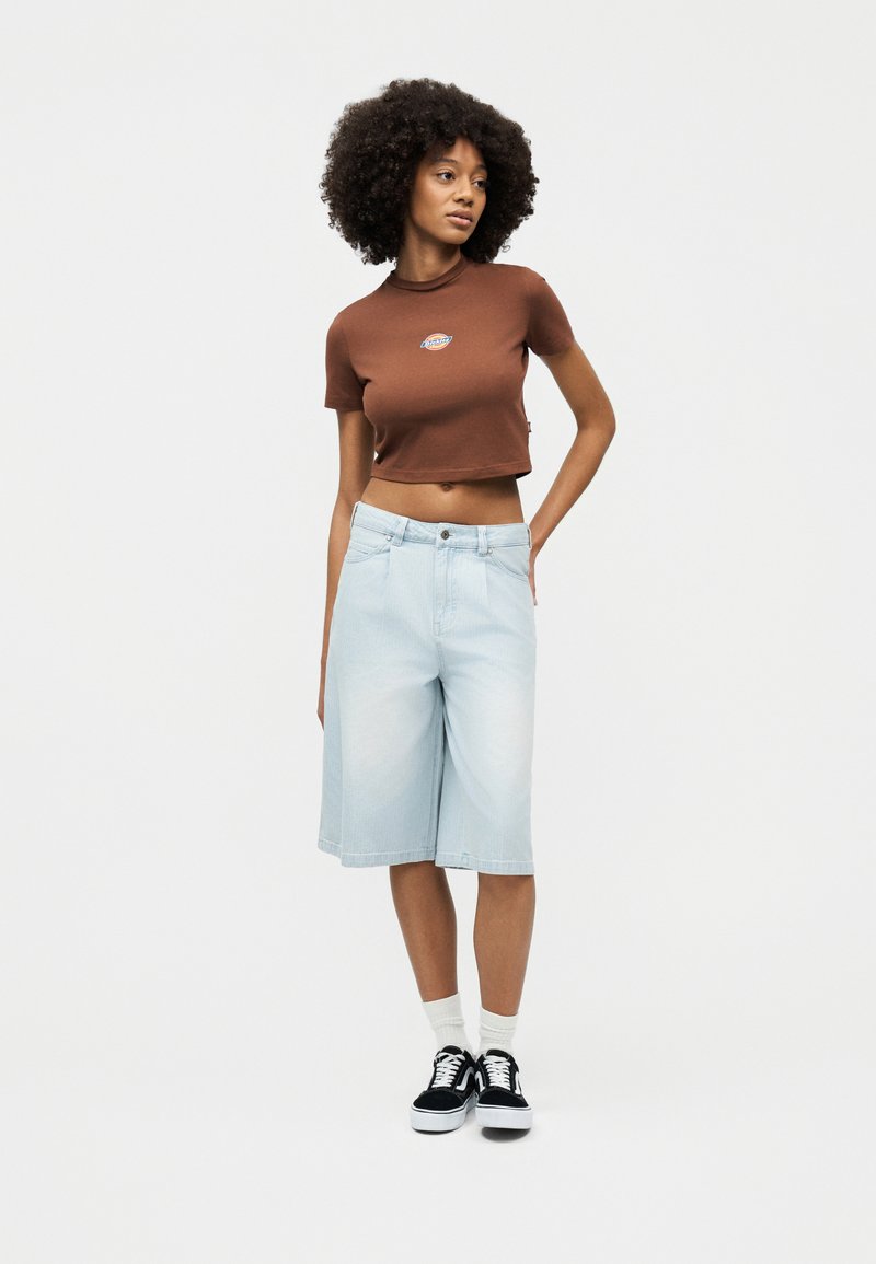 Donna con capelli ricci che indossa una t-shirt marrone corta, pantaloncini di jeans chiari fino al ginocchio, calzini bianchi e scarpe da ginnastica nere, in piedi con una mano sui fianchi.