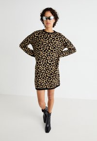 Megzta suknelė su leopardų raštu juodos ir smėlinės spalvų, apvaliu kaklu, ilgomis rankovėmis ir laisvu siluetu, derinama su juodais kulkšnies batais.