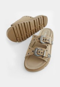 Beige slip-on szandálok, dupla fémbucklival, szegecsekkel díszítve, vastag gumi talppal, amely barázdált talppal rendelkezik a tapadás érdekében.