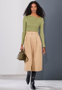 Femme aux longs cheveux bouclés portant un pull côtelé vert, une jupe midi plissée beige, des bottes noires et tenant un sac à main vert olive.