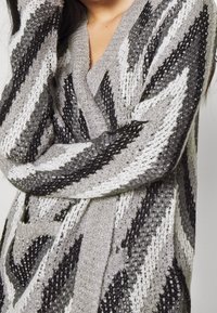 Cardigan en maille épaisse dans des tons gris avec un motif chevron. Manches longues et décolleté en V profond. Tissu doux et texturé.