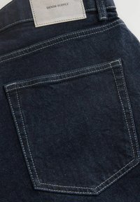Donkerblauwe spijkerbroek met een achterzak, voorzien van contrasterende stiksels en een leren label met de opdruk "DENIM SUPPLY."