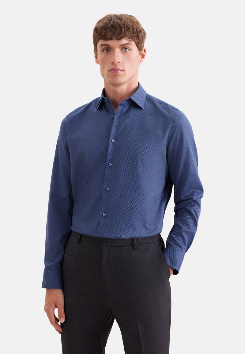 Jeune homme portant une chemise bleue cintrée à boutons avec manches longues et un pantalon gris foncé, debout devant un fond clair uni.