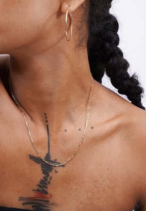 Gold hoop earring and thin gold chain necklace against a tattooed collarbone; skin texture is smooth with visible freckles.