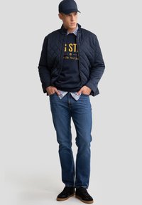 Jūrininkų stiliaus siuvinėta striukė ant tamsaus džemperio su geltonu tekstu, mėlyni džinsai ir juodi sportbačiai. Yra stovinčio apykaklė ir laisvas modelis.