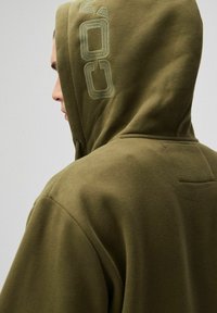 Oliwkowa bluza z kapturem z teksturowanej tkaniny, dużym kapturem i haftowanym logo z tyłu, o gładkim wykończeniu.