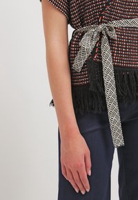 Flerfärgad topp med ett mönstrat tyg i röd, orange och vit, avslutad med en svart frans och ett svartvitt mönstrat bälte.