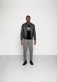 Black leather jacket, black graphic T-shirt, gray tailored trousers, and black lace-up shoes. Model stands centered, neutral background.