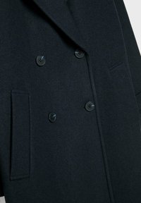 Dvojradový vlnený kabát v tmavomodrej farbe s textúrovaným rybím kosťovým vzorom, vybavený čiernymi gombíkmi a bočnými vreckami.