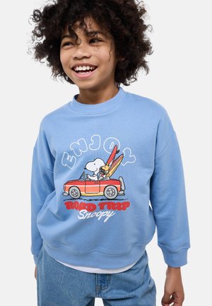 Enfant souriant aux cheveux bouclés portant un sweatshirt bleu avec Snoopy conduisant une voiture rouge chargée de planches de surf, avec le texte « Enjoy Road Trip Snoopy ».