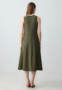Robe sans manches vert olive avec un corsage ajusté, longueur midi évasée et un discret zip au dos. Fabriquée en tissu léger et texturé.