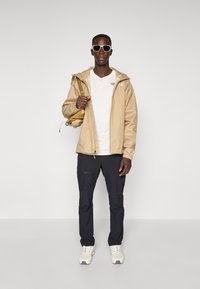 Man wearing beige jacket, white t-shirt, black pants, beige sneakers, sunglasses, and carrying a beige backpack on one shoulder against plain background.