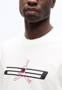 Κοντινό πλάνο ενός ατόμου που φοράει λευκό πουκάμισο με κόκκινο περίγραμμα ενός παίκτη μπάσκετ σε άλμα και δύο οριζόντιες μαύρες ρίγες στο στήθος.