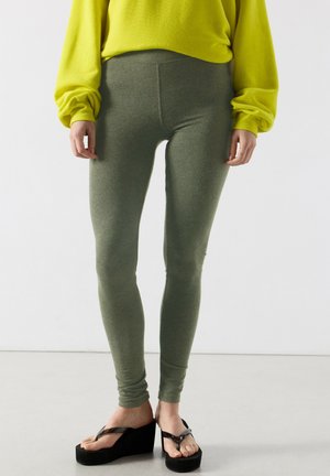 Oseba, ki nosi olivno zelene pajkice, svetlo rumeno majico z dolgimi rokavi in črne platformne japonke, stoji na svetli talni površini.